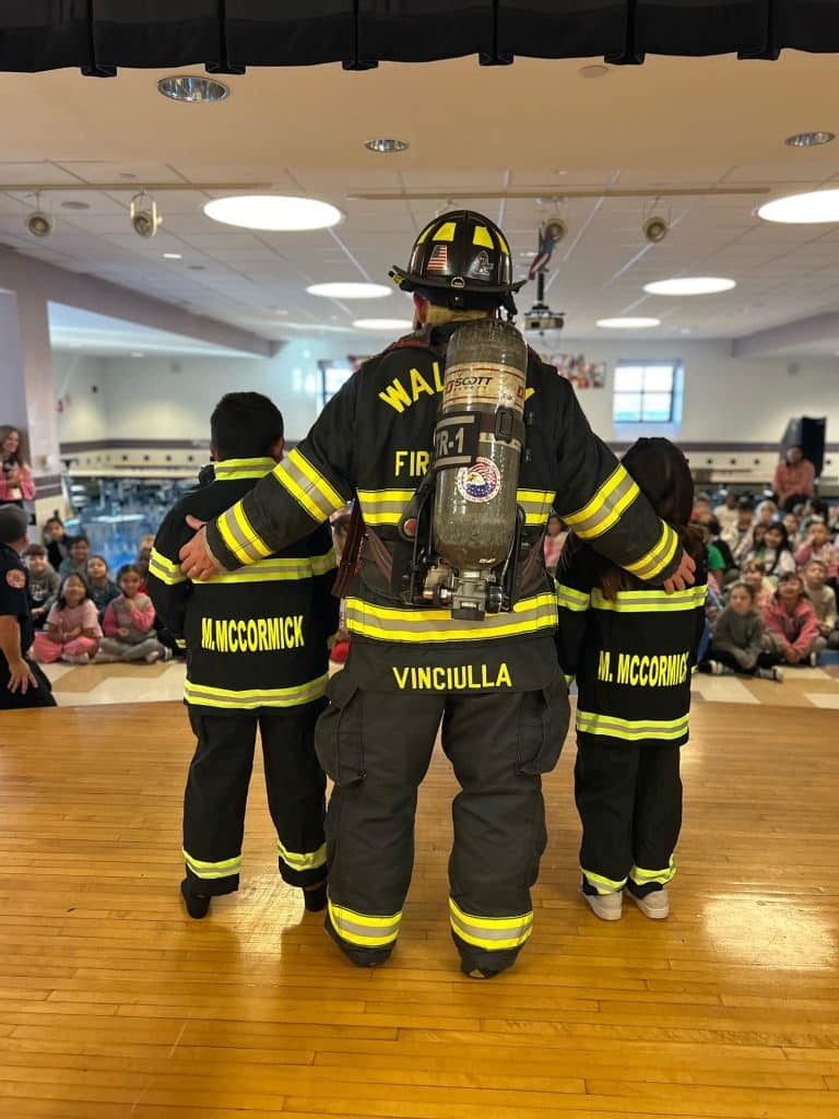 View from behind of Waltham firefighter in full gear flanked buy two children in child-size versions of fire gear.